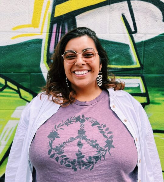 Smiling woman wearing a purple peace sign shirt and white jacket.