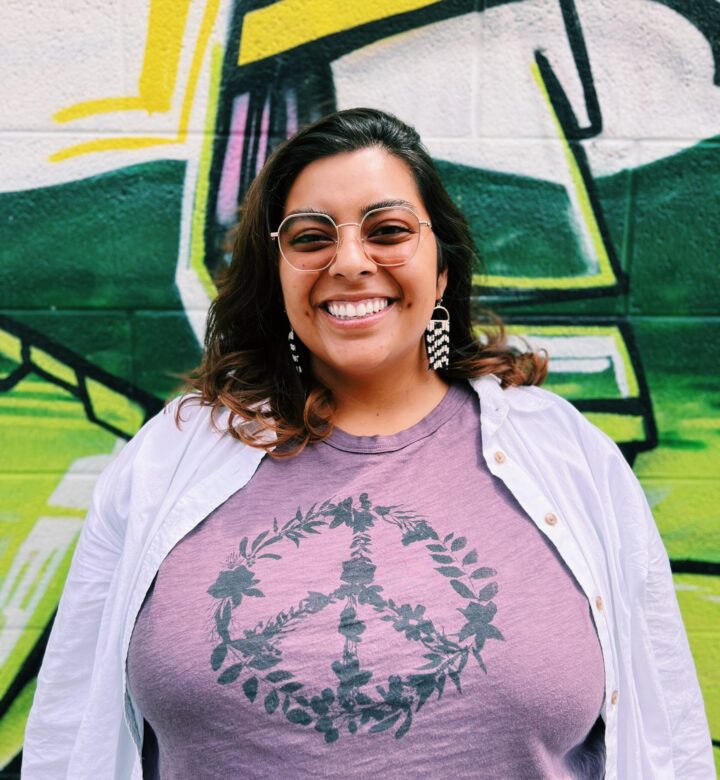 Smiling woman wearing a purple peace sign shirt and white jacket.