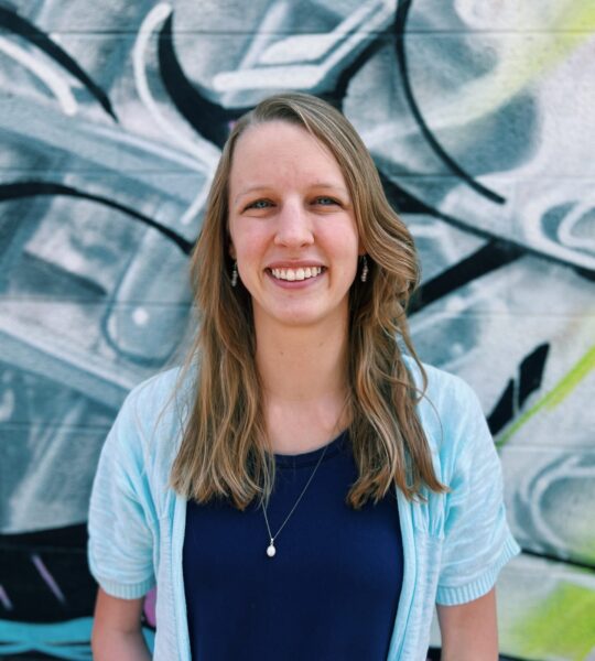 Smiling woman with long hair in casual clothing in front of graffiti.