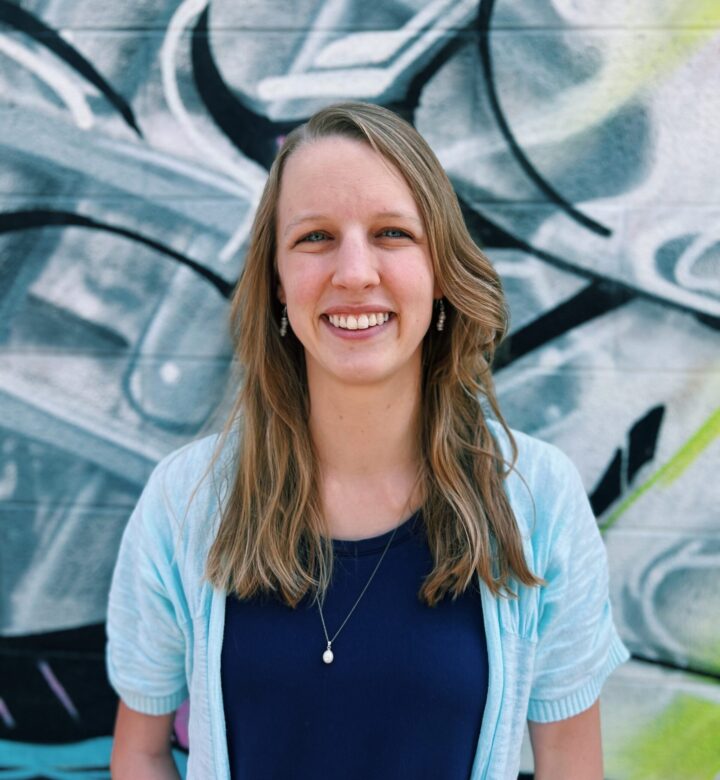 Smiling woman with long hair in casual clothing in front of graffiti.