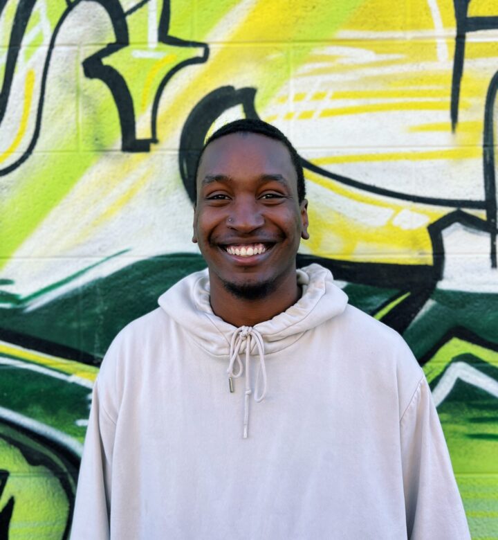 Smiling man in white hoodie stands before a colorful graffiti wall.