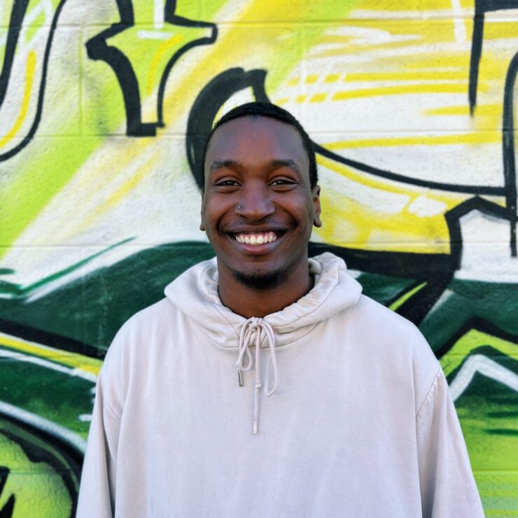 Smiling man in white hoodie stands before a colorful graffiti wall.